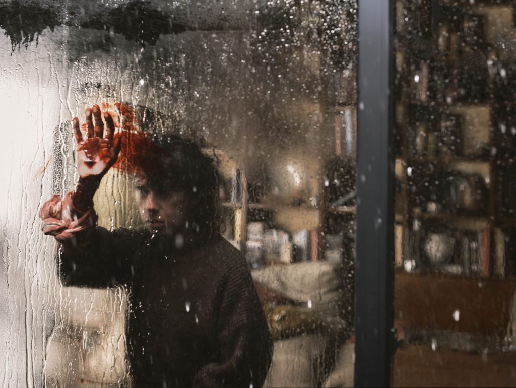 Sally Hawkins as Laura angrily looks out the window at the rain with a bloody hand in Bring Her Back (2025) - Filmotomy
